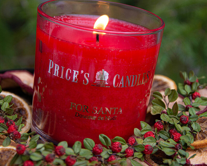 In einer winterlich dekorierten Schale befinden sich einige Zweige mit getrockneten roten Schneeball-Beeren, Zimtstangen sowie getrockneten Apfel- und Orangenscheiben. Dazwischen steht eine Duftkerze im Glas mit dem Namen "For Santa" von Prices Candles. Ihr warmer Kerzenschein schafft eine wohlige und gemütliche Atmosphäre, die an besinnliche Winterabende erinnert.