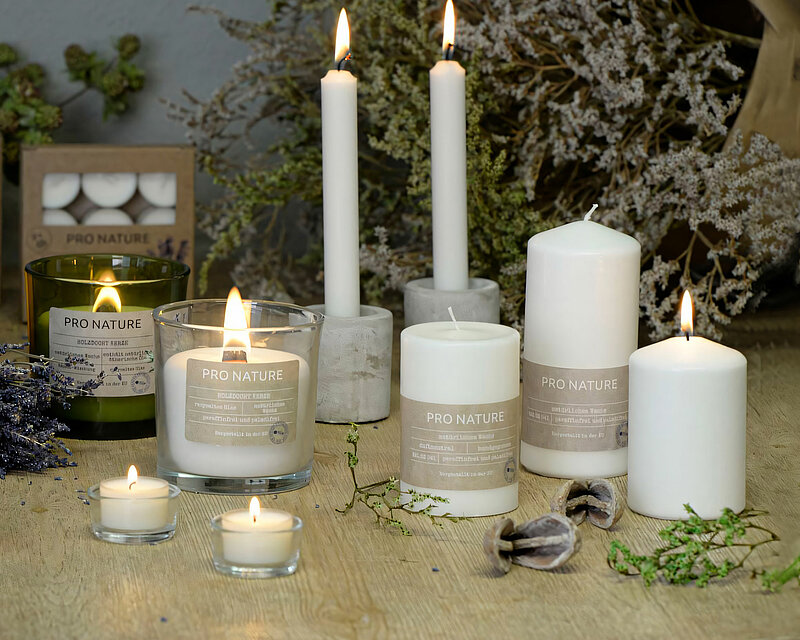 Auf dem Bild befinden sich mehrere Kerzen von der Pro Nature Kollektion von Müller Kerzenfabrik. im Vordergrund zwei brennende Teelichter mit weissem Wachs im durchsichtigen Becher. drei Stumpenkerzen aus weissem Wachs wovon eine brennt. Daneben eine brennende Kerze im Glas mit weisem Wachs und einem Holzdocht. Dahinter stehen zwei graue Beton Kerzenhalter die zwei weisse und brennende Kronenkerzen halten. Daneben eine brennende Duftkerze mit Holzdocht in einem grünen Glas. Dahiner sieht man verschwommen eine braune Packung mit Duftenden Teelichter im durchsichtigen Becher. Alle Artikel stehen auf einem dicken braunen Holztisch. Um die Artikel herum verstreit sind zweige und im Hinergrund ist ein großer Büschel verschiedene trockene zweige. Die Wand im Hintergrund ist leicht verschwommen und grau.