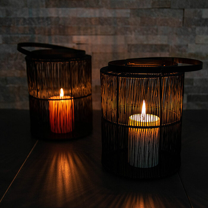 Zwei brennende Kerzen in schwarzen, drahtgeflechtartigen Laternen stehen auf einer dunklen Oberfläche. Die linke Laterne enthält eine orangefarbene Kerze, während die rechte Laterne eine weiße Kerze beherbergt. Der Hintergrund besteht aus einer Wand mit s