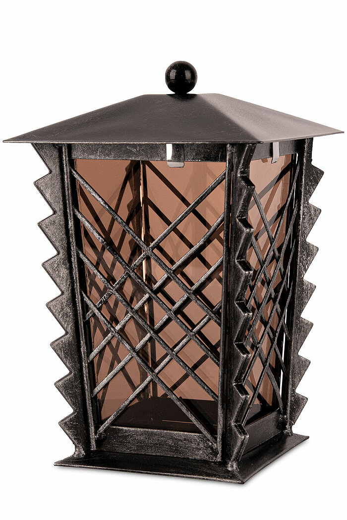 Ein dekoratives, schwarzes Metall-Laternengehäuse mit einem Gitterdesign. Die Laterne hat eine gezackte, diagonale Streifenverzierung an den Ecken und einen spitzen Deckel mit einer kleinen, runden Kugel an der Spitze. Die Seiten bestehen aus transparente