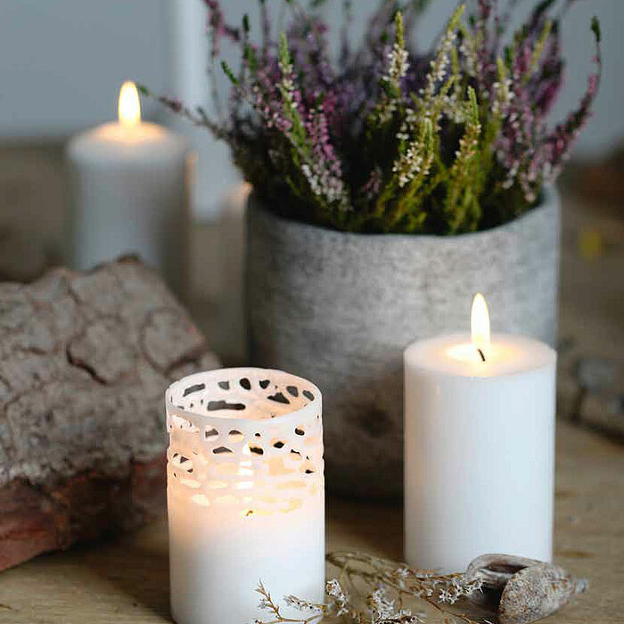 Zwei brennende Müller Stumpenkerzen aus weißem Wachs sind auf einem Holztisch platziert, eine in einem durchbrochenen weißen Kerzenhalter. Dazwischen liegen trockene Zweige und Dekorationsstücke. Dahinter steht eine Betonvase mit buntem Heidekraut. Eine w