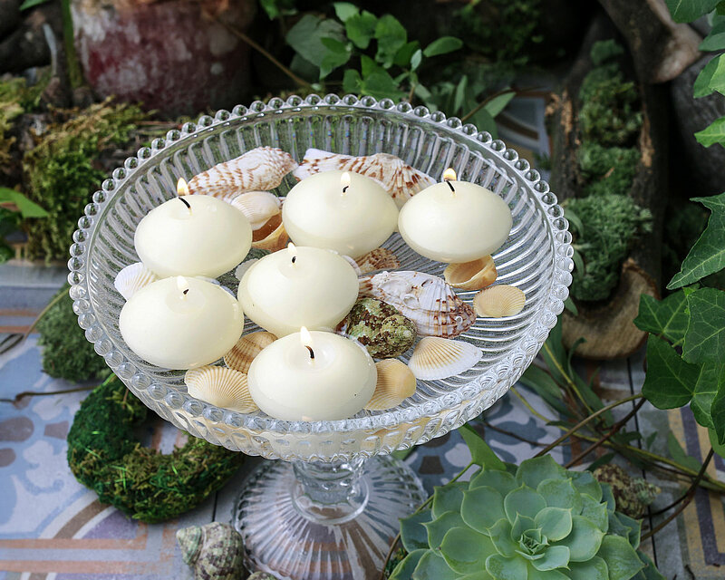Das Bild zeigt eine im Freien aufgenommene Szene. Auf farbenfroh bemalten Terrassenplatten steht eine gläserne Kristallschale, gefüllt mit Wasser, Steinen und Muscheln. Auf der Wasseroberfläche treiben cremefarbene Schwimmkerzen in verschiedenen Größen, die warmes Kerzenlicht verbreiten. Um die Schale herum und im Hintergrund rankt sattes grünes Efeu, während der Boden von weichem Moos bedeckt ist. Die Atmosphäre wirkt friedlich und harmonisch.