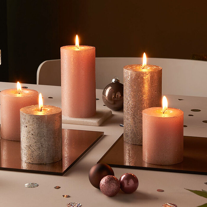 Sechs brennende Bolsius Rustik Shimmer Kerzen in verschiedenen Größen in Rosatönen auf spiegelnden Untersetzern, verteilt auf einem festlich dekorierten Tisch. Ringsherum liegen Glitzerkonfetti und Weihnachtskugeln, im Hintergrund erkennt man einen geschm