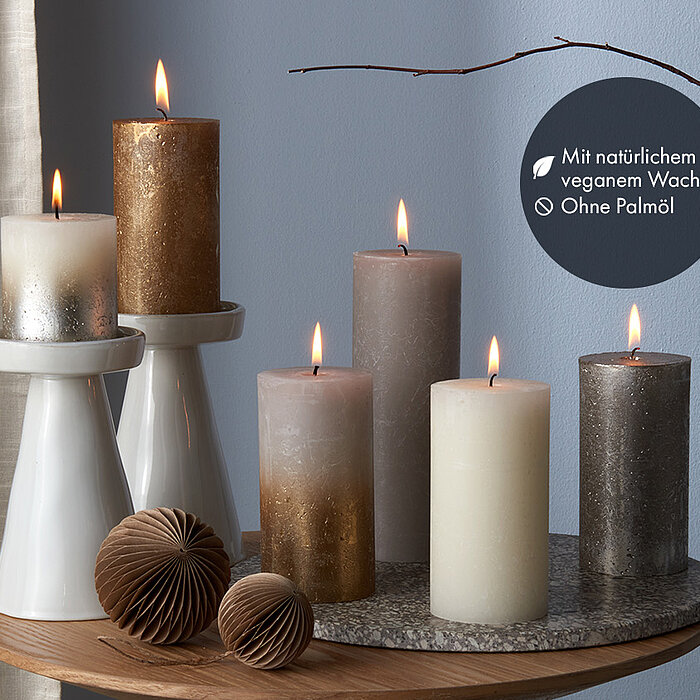 Sechs brennende Bolsius Rustik Shimmer Kerzen in verschiedenen Höhen und Farben, darunter Weiß, Gold, Grau und Silber. Die Kerzen stehen auf weißen Kerzenhaltern und einem runden Holztisch. Zusätzlich sind zwei braune Papierornamente und kahle Zweige als 