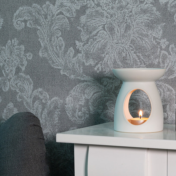 Ein eleganter Raum mit einer brennenden Duftkerze in einem weißen, modernen Keramikhalter, der auf einem weißen Tisch neben einer kleinen Flasche ätherischen Öls steht. Die Wand im Hintergrund ist mit einem grauen, floralen Muster tapeziert und ein dunkel