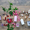 Auf einem rustikalen Holztisch sind verschiedene Kerzen des Herstellers "Müller" in einer Mischung aus Glas- und Stumpenvarianten in den Farben Weiß, Rosa und Rot arrangiert. Dazu sind dekorative Elemente wie Baumwollzweige, Rosen, Beeren und Lavendel ver