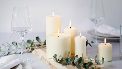 Das Bild ist in hellen und freundlichen cremefarbenen und weißen tönen. Es strahlt eine gemütliche ruhe aus. Auf einem weißen Tisch liegt ein helles Holzbrett mit mehreren grünen Blätterzweigen. Es befinden sich fünf unterschiedlich hohe elfenbeinfarbene brennende Stumpenkerzen nebeneinander und hintereinander welche eine ruhige und gemütliche Atmosphäre ausstrahlen. Am linken Bildrand ist ein schönes kristallklares Weinglas.
