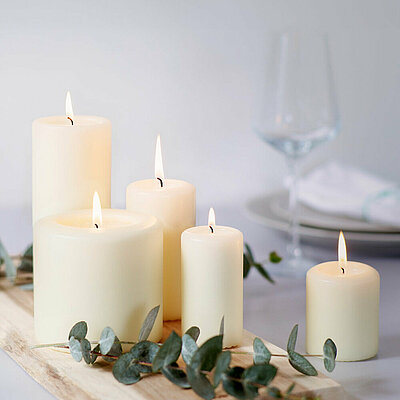 Das Bild ist in hellen und freundlichen cremefarbenen und weißen tönen. Es strahlt eine gemütliche ruhe aus. Auf einem weißen Tisch liegt ein helles Holzbrett mit mehreren grünen Blätterzweigen. Es befinden sich fünf unterschiedlich hohe elfenbeinfarbene brennende Stumpenkerzen nebeneinander und hintereinander welche eine ruhige und gemütliche Atmosphäre ausstrahlen. Am linken Bildrand ist ein schönes kristallklares Weinglas.