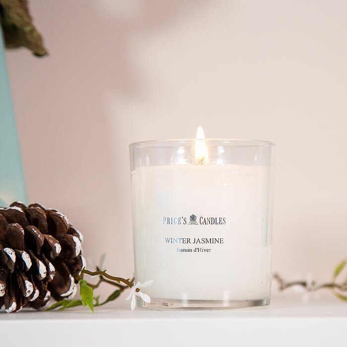 Eine brennende Price's Candles Duftkerze im Glas mit der Aufschrift "Winter Jasmine". Die Kerze steht auf einer weißen Oberfläche, daneben liegt ein verziertes Tannenzapfen und einige grüne Zweige. Im Hintergrund ist ein mintgrünes Dekoelement sichtbar.