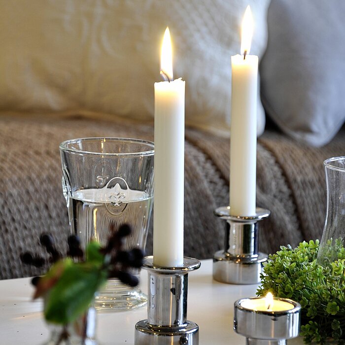 Zwei brennende weiße Stabkerzen von Müller in silberfarbenen Kerzenhaltern und eine brennende weiße Teelichtkerze von Müller in einem passenden Halter stehen auf einem weißen Tisch. Daneben befinden sich ein Wasserglas, eine kleine Vase mit Pflanzen und e
