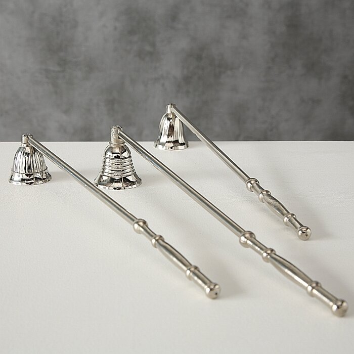 Drei silberne Kerzenlöscher mit unterschiedlichen, kunstvoll gestalteten Glockenköpfen liegen auf einer weißen Oberfläche. Die Hintergrundwand hat eine graue, strukturierte Oberfläche, die einen kontrastreichen Hintergrund bildet.