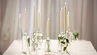 Ein Tisch mit weißer Tischdecke steht vor einem romantischen weißen Vorhang. Auf dem Tisch stehen verschiedene brennende Spitzkerzen und Tafelkerzen in elfenbein. Sie stecken in mit Wasser gefüllten Glasflaschen, in denen verschiedene Grüne Zweige und Blätter schwimmen.