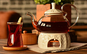 Weißes Keramikstövchen mit einer Kanne Tee darauf, daneben steht ein hohes Glas mit Tee und Löffel. Im Stövchen brennt ein Teelicht mit 4 Stunden Brenndauer.