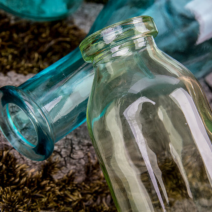 Vier verschiedene Glasflaschen in Gelb, Grün und Türkis in unterschiedlichen Größen von "Sandra Rich" liegen auf einem Stück Baumrinde. Die Flaschen haben eine rustikale, antike Ästhetik und sind stimmungsvoll im Freien inszeniert.