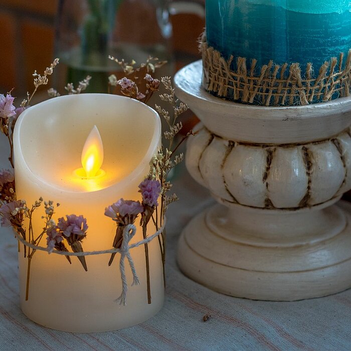 Eine weiße LED-Kerze mit realistisch flackernder Flamme steht auf einem Tisch. Die Kerze ist mit einem filigranen Kranz aus getrockneten Blumen und Zweigen dekoriert. Rechts daneben steht eine größere, blaue Stumpenkerze auf einem dekorativen, weißen Kerz
