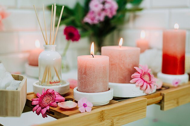 In einem romantischen Badezimmer steht auf dem Badewannenrand eine Badewannenablage aus Holz. Auf der Ablage liegen schöne pinke Blumenblüten und einige unterschiedlich farbige Bolsius Rustik Kerzen in Rot- und Rosatönen. Dahinter steht ein Bolsius Raumduft-Diffuser mit Holzstäbchen, der einen angenehmen Duft verströmt. Im Hintergrund ist verschwommen eine Vase mit saftig grünen, leuchtenden Pfingstrosen zu sehen. Die Szene strahlt eine beruhigende und romantische Atmosphäre aus, die zum Entspannen und Genießen einlädt.