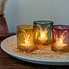 Drei brennende Teelichter in dekorativen Glasbehältern in den Farben Gelb, Grün und Rosa vom Hersteller "Wurm". Die Gläser stehen auf einem runden, weißen Keramikteller, der mit Strohelementen verziert ist. Im Hintergrund befindet sich eine Vase mit getro