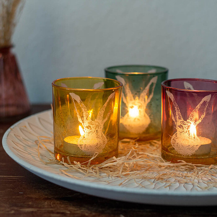 Drei brennende Teelichter in dekorativen Glasbehältern in den Farben Gelb, Grün und Rosa vom Hersteller "Wurm". Die Gläser stehen auf einem runden, weißen Keramikteller, der mit Strohelementen verziert ist. Im Hintergrund befindet sich eine Vase mit getro