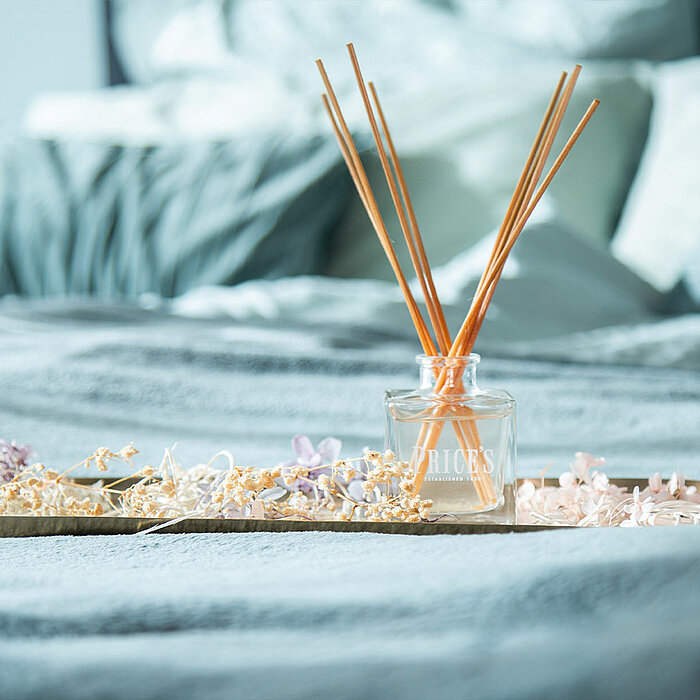 Ein goldfarbenes Tablett auf einem grauen Bett. Auf dem Tablett stehen mehrere Holzstäbchen in einem Glasbehälter von "Prices Candles". Um den Behälter herum sind getrocknete Blumen in Rosa und Beige arrangiert. Der Hintergrund zeigt ein gemütliches Bett 