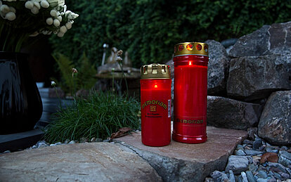 zwei verschieden hohe rote Grabkerzen Memoriam von Aeterna mit Deckel stehen auf einem Stein vor einer Steinwand. Im Hintergrund ist eine grüne Hecke und einige weisse Blumen.