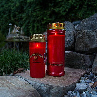zwei verschieden hohe rote Grabkerzen Memoriam von Aeterna mit Deckel stehen auf einem Stein vor einer Steinwand. Im Hintergrund ist eine grüne Hecke und einige weisse Blumen.