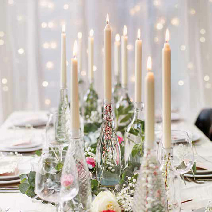 Elf brennende Bolsius Tafelkerzen in elegant arrangierten Glasflaschen auf einem festlich gedeckten Tisch. Die Tischdekoration umfasst grüne Blätter, weiße und rosa Blumen sowie zarte Glaswaren, die in einem filigranen Zusammenspiel eine romantische Atmos