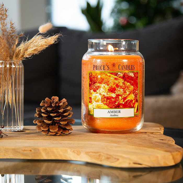 Eine brennende Prices Candles Kerze "Oriental Nights" steht auf einem Tisch in einem modernen Wohnzimmer. Auf dem Tisch befinden sich außerdem zwei goldene Behälter auf einem weißen Tablett, eine kleine Buddha-Statue und eine Vase mit gelben Blumen. Im Hi