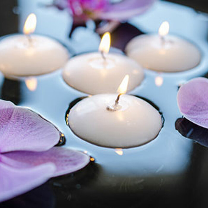 Fünf brennende weiße Schwimmkerzen treiben auf einer Wasseroberfläche, umgeben von zartrosa Orchideenblüten. Das Bild strahlt eine ruhige und entspannende Atmosphäre aus.