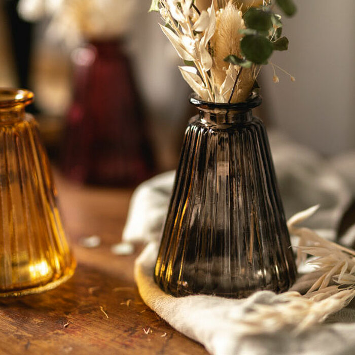 Drei geriffelte Glasvasen von Sandra Rich in Bernstein, Rauchgrau und Smaragdgrün stehen auf einem hölzernen Tisch. Die graue Vase ist mit getrockneten Gräsern gefüllt. Eine rote Vase steht unscharf im Hintergrund. Alles ist in warmes, weiches Licht getau
