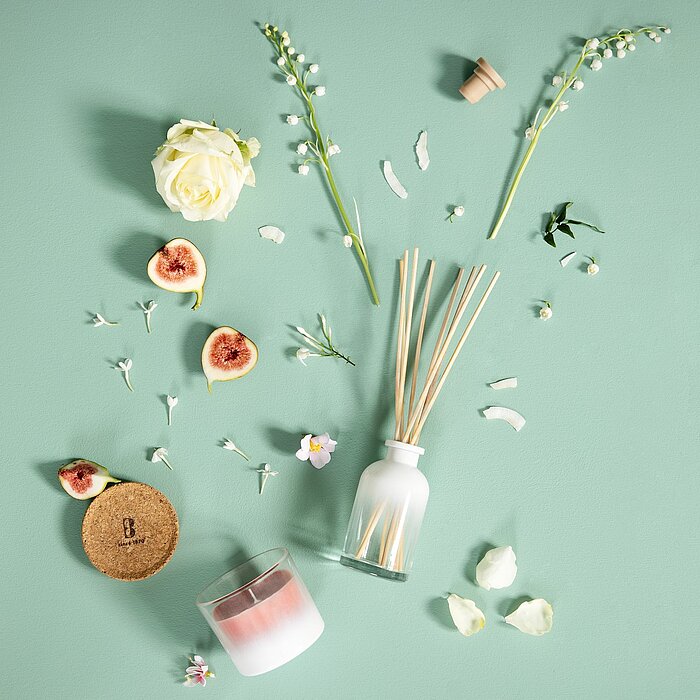 Eine brennende Bolsius Duftkerze in einem halbtransparenten Glasbehälter. Die Kerze steht auf einem Holz-Tisch, daneben liegen Schmuckstücke und eine Schere auf einer Muschelschale. Rechts daneben steht eine Glasflasche mit Duftstäbchen. Im Hintergrund si