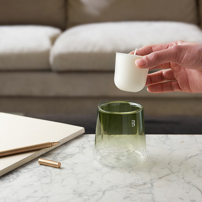 Eine Hand setzt ein weißes Teelicht in ein grünes Glasgefäß auf einem Marmortisch. Daneben liegen ein Notizbuch und ein goldener Stift mit abgenommenem Deckel. Im Hintergrund steht ein beiges Sofa mit weichen Kissen, was eine gemütliche Atmosphäre erzeugt