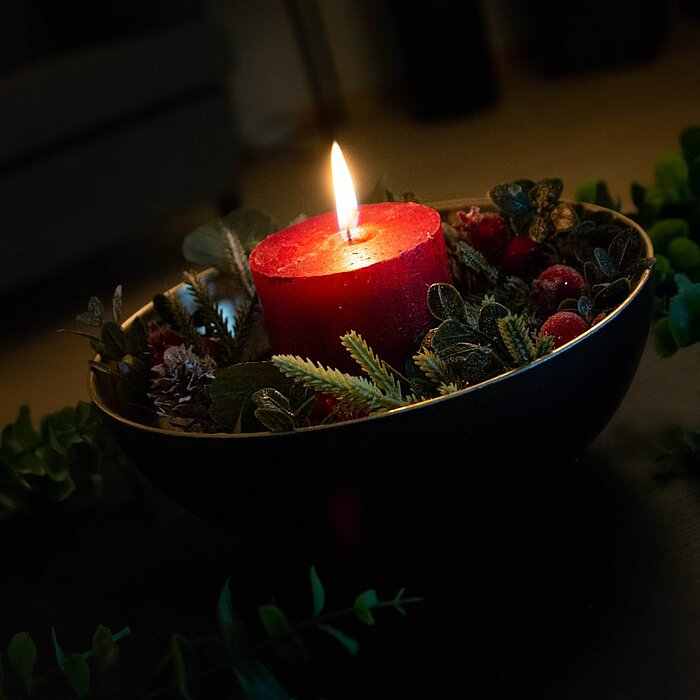 Eine einzelne brennende rote Kerze steht in der Mitte einer goldenen Schale, die mit Tannenzweigen, Zapfen und roten Beeren dekoriert ist. Die gesamte Komposition verleiht dem Bild eine warme und festliche Atmosphäre. Um die Schale herum sind verstreut gr
