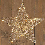 Ein leuchtender Stern aus filigranem Drahtgeflecht, der mit kleinen warmweißen Lichtern durchzogen ist, hängt vor einer rustikalen Holzwand. Die Lichter verleihen dem Stern eine gemütliche und festliche Ausstrahlung.