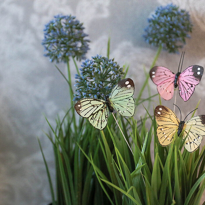 Das Bild zeigt filigrane, bunte Schmetterlingsdekorationen vom Hersteller Steingaesser, die an hohen, grünen Kunstpflanzen angebracht sind. Im Hintergrund sind zarte, lilafarbene Blüten und eine florale Tapete zu sehen.