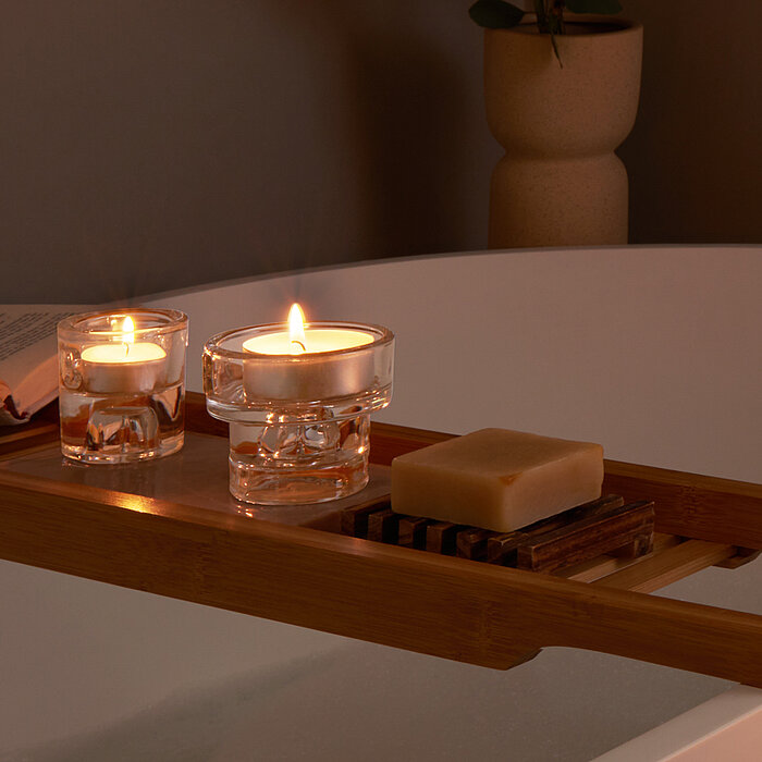Zwei brennende Bolsius Teelichter in Glasbehältern stehen auf einem hölzernen Badewannen-Tablett. Neben den Teelichtern liegen ein offenes Buch und ein Stück Seife auf einer Holzablage. Die Szene erzeugt eine entspannende und behagliche Atmosphäre.