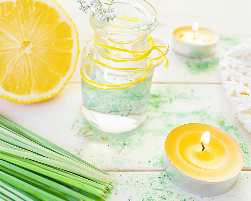 Ein frisches Bild mit einem weißen Tisch, auf dem aufgeschnittene Zitronen, Zitronengras und eine Glaskaraffe mit Wasser stehen. Daneben brennen zwei gelbe Citronella Duft-Teelichter.