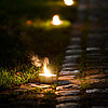 Mehrere brennende "Müller" Teelichter säumen einen steinernen Weg, der durch eine grasbewachsene Fläche führt. Die warmen Lichter der Teelichter erzeugen eine gemütliche und einladende Atmosphäre in der abendlichen Dämmerung.