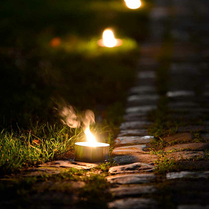 Mehrere brennende "Müller" Teelichter säumen einen steinernen Weg, der durch eine grasbewachsene Fläche führt. Die warmen Lichter der Teelichter erzeugen eine gemütliche und einladende Atmosphäre in der abendlichen Dämmerung.