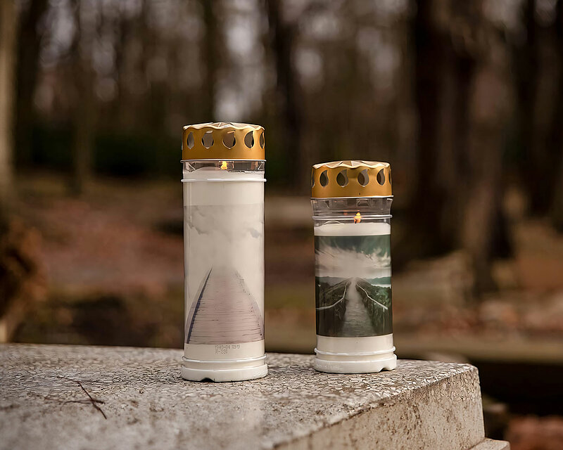 Eine große und eine kleine brennende Grabkerze mit Deckel und Motiv von Bolsius stehen nebeneinander auf einem grau marmorierten Grabstein. Auf dem Rest des Bildes sieht man einen herbstlichen Wald und weitere Grabsteine.