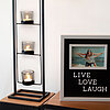 Drei brennende Teelichter in Glasbehältern sind in einem Metallgestell vertikal angeordnet. Daneben steht ein gerahmtes Schild mit der Aufschrift "LIVE LOVE LAUGH" und einem eingerahmten Foto von zwei Personen am Strand. Die Szenerie strahlt eine gemütlic