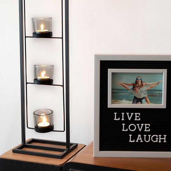 Drei brennende Teelichter in Glasbehältern sind in einem Metallgestell vertikal angeordnet. Daneben steht ein gerahmtes Schild mit der Aufschrift "LIVE LOVE LAUGH" und einem eingerahmten Foto von zwei Personen am Strand. Die Szenerie strahlt eine gemütlic