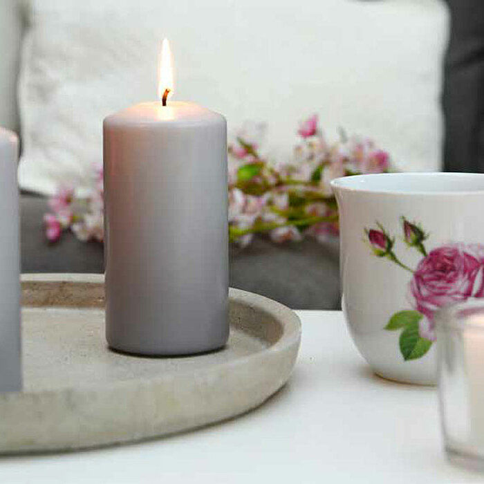 Zwei brennende weiße Müller Kerzen sind in transparenten Glasbehältern auf einer strukturierten grauen Unterlage platziert. Eine große rosa Blüte und eine Muschel liegen neben den Kerzen. Im Hintergrund sind grüne Pflanzen und weitere rosa Blüten zu sehen