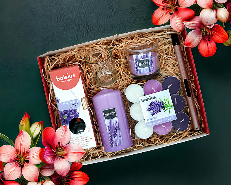 Man blickt von oben auf eine DecoLite Geschenkebox die geöffnet ist. In der Box befindet sich etwas Holzwolle und einige Artikel unterschiedlicher Hersteller mit dem Duft Lavendel. Um die Box herum sind einige rosa und rote Farbene Blüten und knospen zu sehen. Der Hintergrund ist in einem schlichten und dunklen grün gehalten sodass die Geschenkebox ins Auge sticht.
