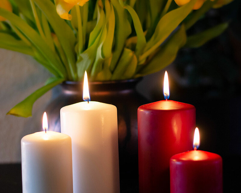 Das Bild hat durch den warmen Kerzenschein eine ruhige und angenehme Ausstrahlung. In der Mitte des Bildes stehen je zwei weiße und zwei rote DecoLite Safe Candle Stumpenkerzen. Die im Hintergrund stehenden Narzissen in einer Vase werden sanft vom Kerzenlicht angestrahlt was eine angenehme und warme Stimmung verbreitet.