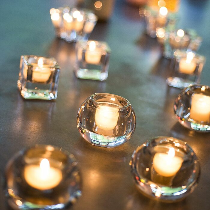 Mehrere brennende Teelichter in transparenten Glasbehältern des Herstellers "Sandra Rich" verteilen sich kunstvoll auf einer glatten Oberfläche. Unterschiedlich große und geformte Halter, teils rund, teils eckig, erzeugen ein sanftes, warmes Licht und ein