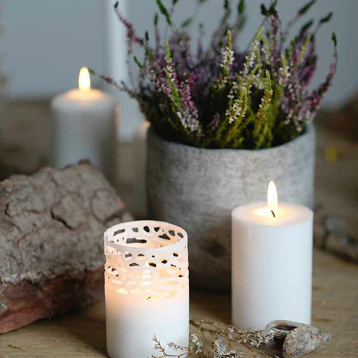 Auf einem Holztisch stehen mehrere brennende weiße Teelichter von Müller. Daneben befinden sich zwei brennende weiße Stumpenkerzen von Müller in unterschiedlichen Größen und ein schwarzes, facettiertes Glas, das mit weißen Stabkerzen von Müller gefüllt is