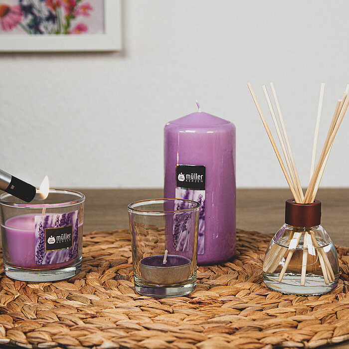 Ein kleines Arrangement von Produkten von "DecoLite" auf einem geflochtenen Tischset. Eine violette Stumpenkerze, ein Glas mit einer violetten Kerze und ein leeres Glas stehen nebeneinander. Daneben ist ein Duftdiffusor mit mehreren Holzstäbchen positioni
