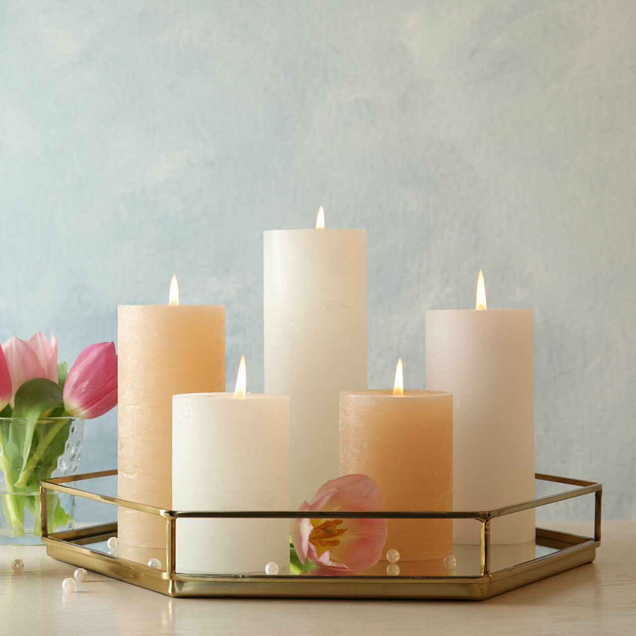 Heller, freundlicher Raum, dekoriert mit verschiedenen Rustik Stumpenkerzen in den Farben weiß und creme. Im Hintergrund ist eine Vase mit rosa Tulpen zu erkennnen.