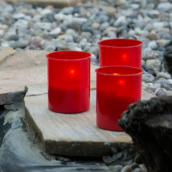 Drei rote brennende Grableuchten vom Hersteller "Müller" stehen auf einer flachen Steinplatte am Rand eines kleinen Teichs. Um die Platte herum sind zahlreiche kleine Kieselsteine verteilt, und im Vordergrund ist ein Teil eines dunklen Baumstamms zu sehen