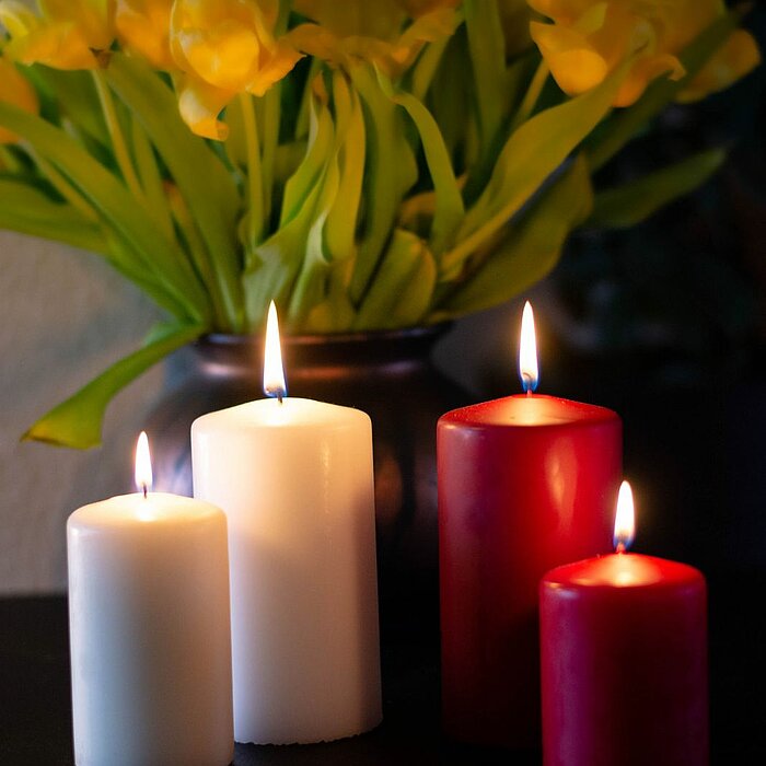 Vier brennende DecoLite Stumpenkerzen in den Farben Weiß und Rot stehen auf einem dunklen Tisch. Im Hintergrund steht eine Vase mit strahlend gelben Tulpen.